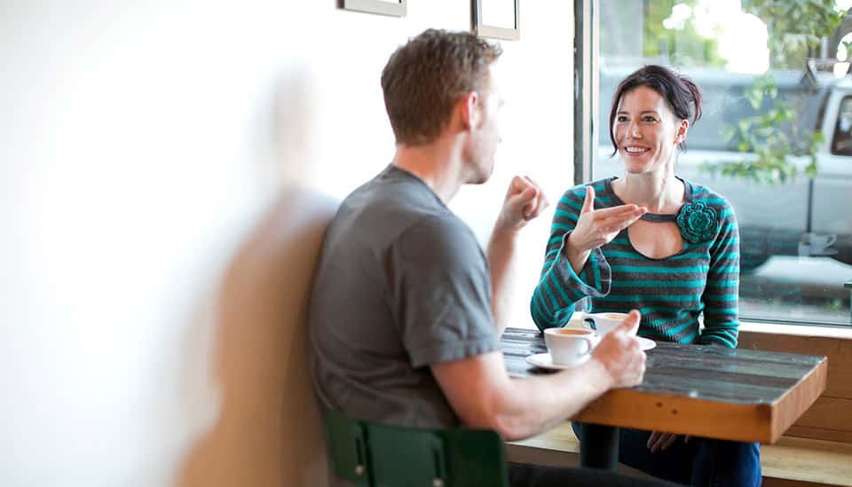Two people sitting at a table in a cafe, engaged in conversation over coffee. One person is smiling and gesturing while the other is listening attentively.