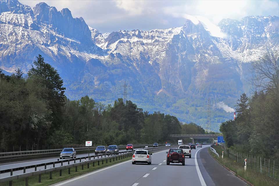 A scenic highway with vehicles traveling through a mountainous landscape, featuring snow-capped peaks and greenery along the road.