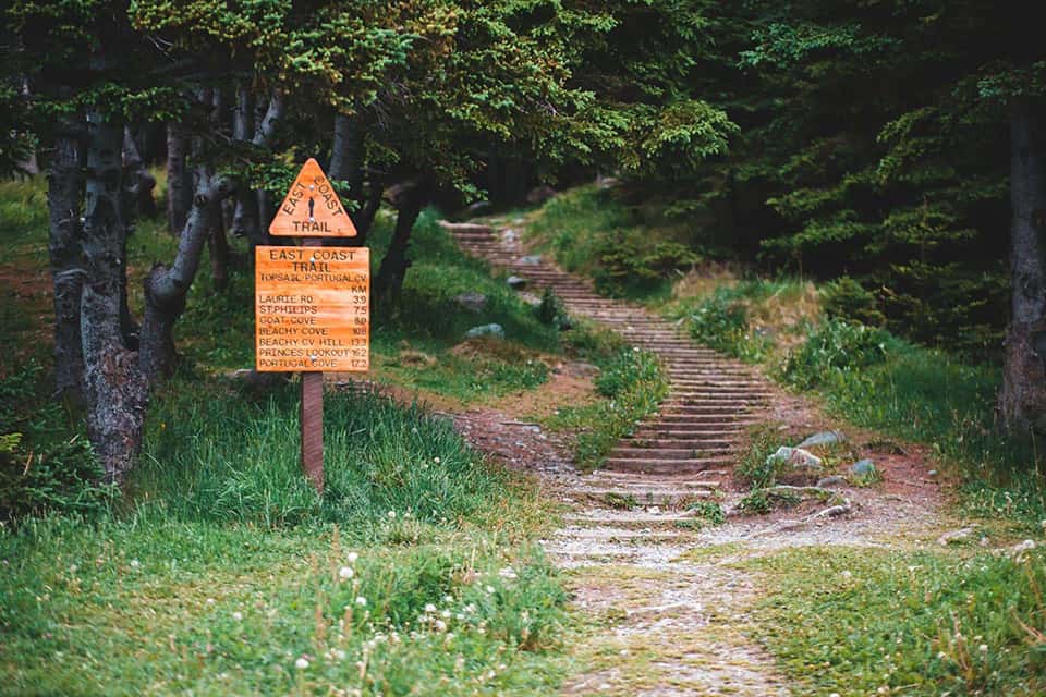 A wooden sign indicating a hiking trail, with information about the path and distances to various destinations. The trail is surrounded by greenery and trees.
